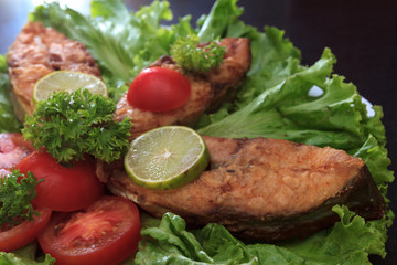 A piece of fried fish fillett served on blue glass table with lettuce, lime, tomato and herbs