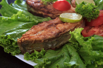 A piece of fried fish fillett served on blue glass table with lettuce, lime, tomato and herbs