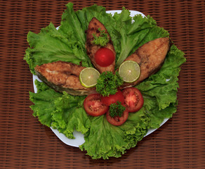 A piece of fried fish fillett served on blue glass table with lettuce, lime, tomato and herbs