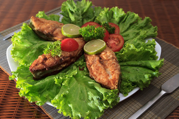 A piece of fried fish fillett served on blue glass table with lettuce, lime, tomato and herbs