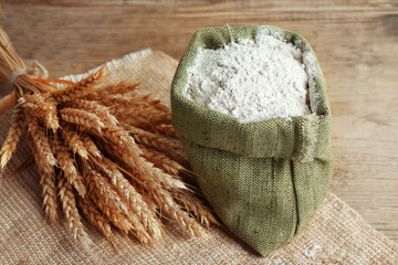 Whole flour in burlap bag with wheat ears on wooden table, closeup