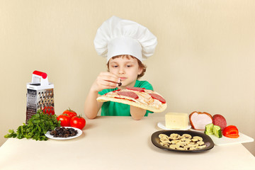 Little boy in chefs hat puts olives on the pizza crust