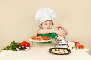 Little funny boy in chefs hat rubs on a grater cheese for pizza