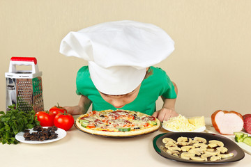 Little boy in chefs hat sniffing the aroma of cooked pizza
