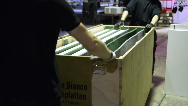Two black dressed men pushing a wooden case of equipment through a hall.