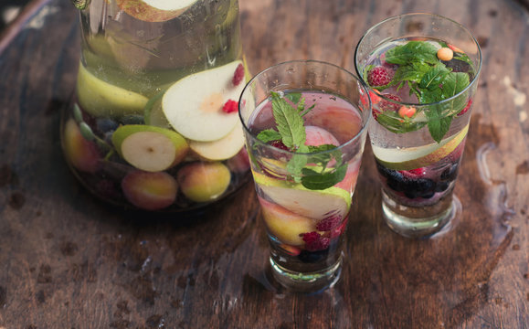 Two glasses of soft drink with mint with seasonal fruit are on a wooden surface on a table in a garden next to a glass beaker with a drink of vegetarian food
