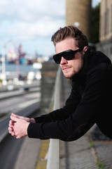 A young and attractive man in his 20s standing outside, wearing a black hoodie and sunglasses, looking away from camera.