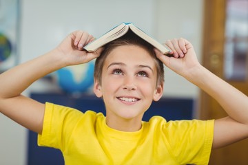 Smiling pupil holding book on head in a classroom