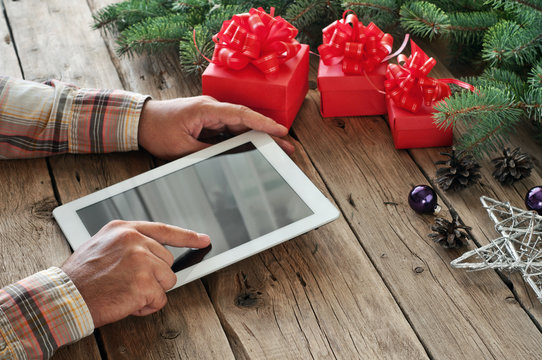 Tablet computer in men hands. Near the table with tablet computer three gift boxes, christmas toys, stars , fir cones and fluffy fir branches. Free space for text. top view. Copy space. Closeup