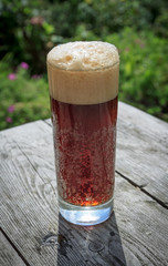 Tall glass full of frothy dark beer on rustic wooden table in summer garden