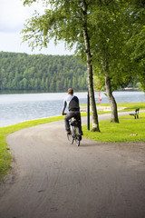 A man riding a bike in the park next to a lake. Some forest is on the other side of the lake and the curve is turning right.