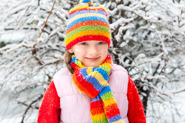 Winter portrait of a cute little girl under the snowfall, wearing red pullover, colorful hat and scarf