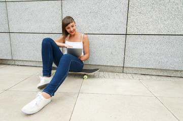 Young girl siting on skateboard in the city with tablet by the w