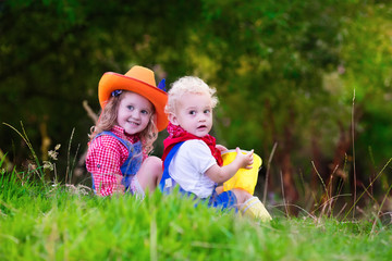 Little boy and girl dressed up as cowboy and cowgirl playing with toy rocking horse in park. Kids play outdoors. Children in Halloween costumes at trick or treat. Toys for preschooler or toddler child