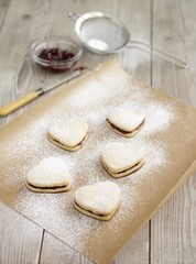 Heart-shaped biscuits filled with jam and dusted with icing sugar on baking paper