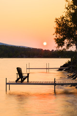 Adirondack chair on the end of a dock on a lake at sunset.