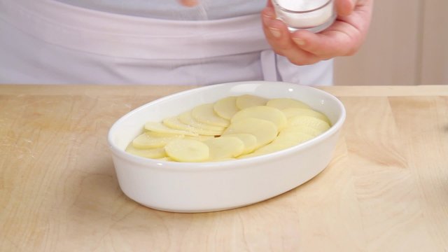 Potato slices in a baking dish being seasoned with salt and pepper