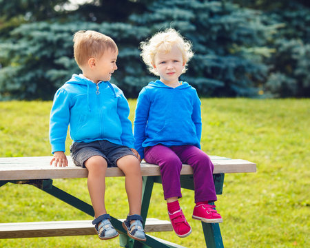 Portraits Of Caucasian Cute Boy And Girl Friends Sitting Together In Park Outside On Summer Day, Backlit With Sun From Behind