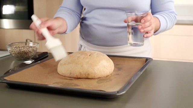 A loaf of bread being brushed with water, scored and sprinkled with sunflower seeds