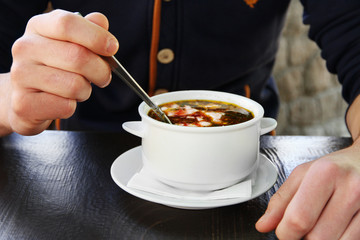 White bowl with borsch and male hands with spoon on wooden table, closeup