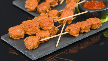 Spicy Chicken Satay - Marinated chicken meatball skewers served with chilli sauce and lemon wedges on a slate with reflections.
