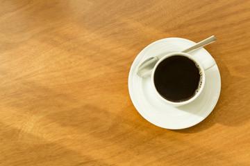 Single black cup of hot coffee in a white mug on a white saucer with a silver spoon on a wooden table in sunlight