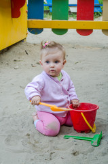 Fototapeta premium Little beautiful girl with a baby barrette color play in the playground sandbox sand pail, shovel and rake