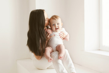 Mother kissing baby in white room at home near window