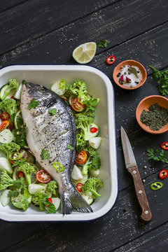 raw ingredients - Dorado fish and vegetables - broccoli, zucchini, onions, peppers, lime and spices on a dark wooden surface, cooking baked fish with vegetables