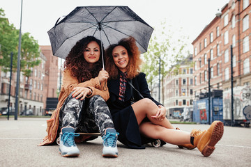 Two young friends sitting on a skateboard with an umbrella