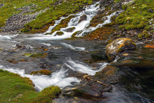 Mountain stream and meadow.
Flowing mountain stream of river with several creeks effluent from out of ground grassy meadow along shore orange red color stones cleaving water
