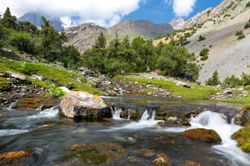 Fototapeta na wymiar Fast mountain creek Rapid water stream of river on foreground and group of people hiker walking up hill footpath on background