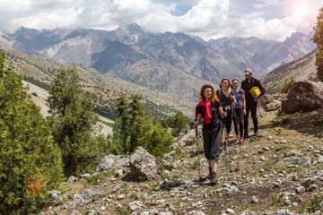 Fototapeta premium Group of people walking on trail Men and women going up with backpack luggage and hiking gear on bright mountain landscape background with sun rising and high peaks behind