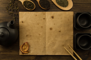 Black teapot, two cups, a collection of tea, dried apples, old blank open book on wooden background. Menu, recipe, mock up