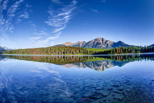 Colorful trees lined the shores of Patricia Lake at Jasper National Park with Pyramid Mountain in the background. The calm lake reflects a mirror image of the mountains and trees.