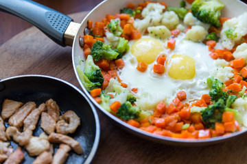 Scrambled eggs with broccoli and carrots. Slices of fried meat in a frying pan