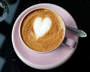 Pink cup of cappuccino, with milk foam in form of heart, on the table in the cafe.