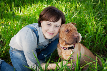 smiling happy young woman in denim overalls hugging her red cute dog Shar Pei in the green grass in park, true friends forever, people concept