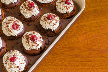 many freshly baked chocolate muffins with whipped cream and cherries on top in a baking form on a wooden table