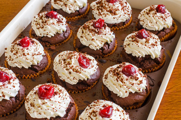 many freshly baked chocolate muffins with whipped cream and cherries on top in a baking form on a table