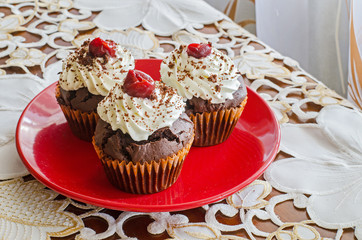 three chocolate muffins with whipped cream and cherry on top on a red plate on a white decorated table cloth 