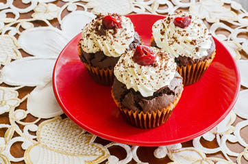 side view of three chocolate muffins with whipped cream and cherry on top on a red plate on a white decorated table cloth 