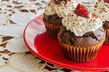 close-up of three chocolate muffins with whipped cream and cherry on top on a red plate on a white decorated table cloth 