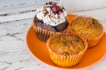 close-up of an orange plate with two lemon muffins and a chocolate muffin with whipped cream and cherry on an old white wooden surface 