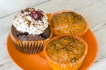 close-up of an orange plate with two lemon muffins and a chocolate muffin with whipped cream and cherry on an old white wooden surface 