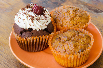 close-up of an orange plate with two lemon muffins and a chocolate muffin with whipped cream and cherry on an old wooden table