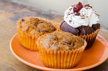 orange plate with two lemon muffins and a chocolate muffin with whipped cream and cherry on an old...