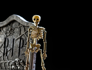 A plastic Halloween skeleton stands next to a carved tombstone against black background.