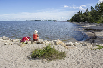 horizontal image of a woman sitting on a large rock on a sandy beach with a red umbrella lying by her side gazing out across the lake on a warm summer day