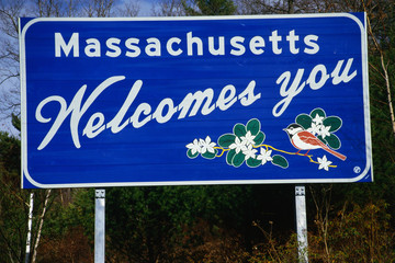 This is a road sign that says Massachusetts welcomes you. It is against a blue sky. On the sign are...